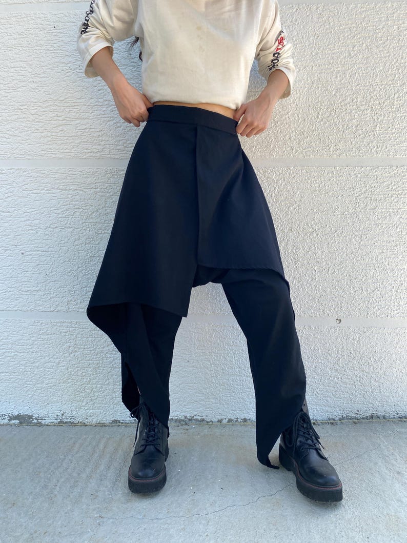 May include: Black trousers featuring an asymmetrical wrap-style skirt overlay that drapes down the front. The garment has a high waistband and wide-leg cut, paired with black lace-up boots. The fabric appears solid and opaque in texture.