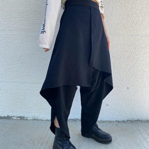 May include: Black trousers featuring an asymmetrical wrap-style skirt overlay that drapes over the legs. The garment has a high waistband and a loose, flowing silhouette that extends to the lower calf, paired with black lace-up platform boots.