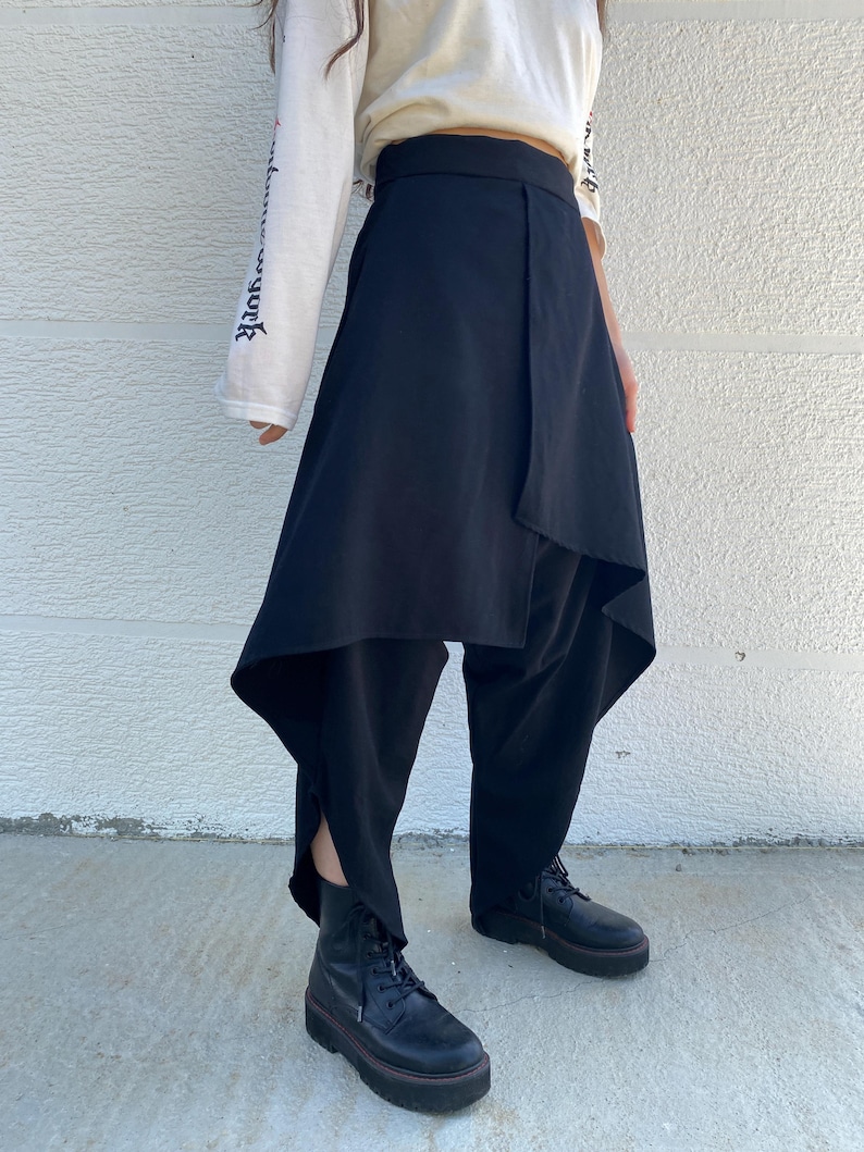 May include: Black trousers featuring an asymmetrical wrap-style skirt overlay that drapes over the legs. The garment has a high waistband and a loose, flowing silhouette that extends to the lower calf, paired with black lace-up platform boots.