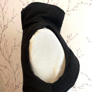Pode incluir: Pe&ccedil;a de vestu&aacute;rio preta sem mangas com gola alta e texturizada. A gola e as bordas apresentam um tecido tecido, enquanto o corpo parece ser um material liso e fosco. A pe&ccedil;a &eacute; exibida em um manequim branco sobre um fundo floral.