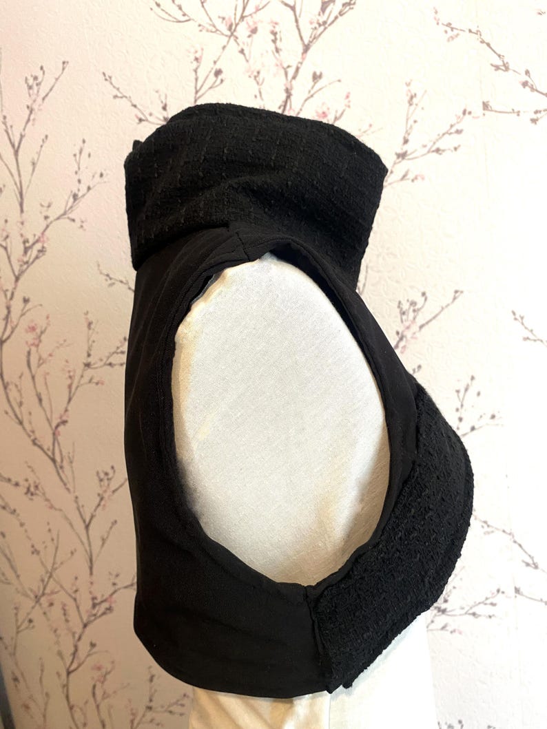 Pode incluir: Pe&ccedil;a de vestu&aacute;rio preta sem mangas com gola alta e texturizada. A gola e as bordas apresentam um tecido tecido, enquanto o corpo parece ser um material liso e fosco. A pe&ccedil;a &eacute; exibida em um manequim branco sobre um fundo floral.