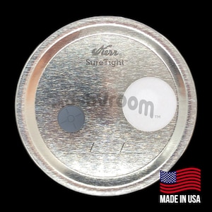 Puede incluir: Tapa de metal plateado con un sello gris y una pegatina blanca con el texto "Kerr Sure Tight" y "oom™". La tapa también tiene el texto "MADE IN USA" impreso en ella.