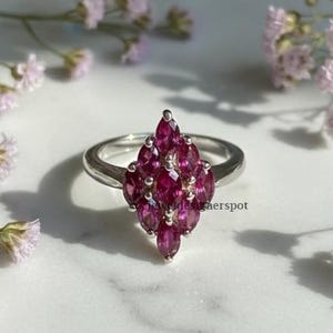 May include: A silver ring featuring a cluster of marquise-cut, deep red gemstones. The gemstones are arranged in a pointed, fan-like design. The ring is set against a backdrop of white marble and delicate purple flowers.