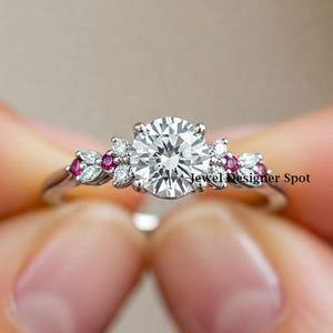 May include: A silver engagement ring with a large round center stone and smaller stones set in a floral design. The ring has small pink stones set in the floral design.
