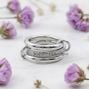 May include: A silver ring with multiple bands, one encrusted with small, clear stones. The ring has a unique design with interlocking loops. The background features small purple flowers and delicate stems, creating a soft, elegant aesthetic.