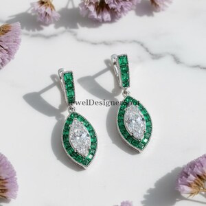 May include: Silver earrings featuring a clear, marquise-cut central stone, framed by emerald-green stones. The earrings are set on a white marble surface, with purple flowers. The earrings have a rectangular top. The earrings are a beautiful piece of jewelry.