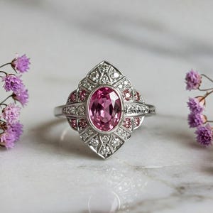 May include: A delicate, oval-shaped pink sapphire engagement ring set in a white gold band. The ring features a vintage-inspired design with a halo of smaller diamonds and pink sapphires.