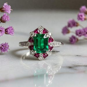 May include: A silver ring with a large emerald green gemstone surrounded by smaller ruby red gemstones and diamonds. The ring is on a white marble surface.