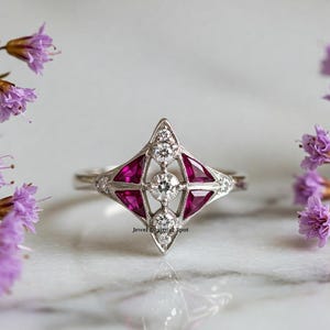 Op de afbeelding: Een zilveren ring met een geometrisch ontwerp met een grote centrale diamant en vier kleinere robijnen. De ring ligt op een witte achtergrond.