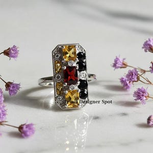 May include: A silver ring with a rectangular setting featuring a mix of yellow, red, and black gemstones. The ring is set against a white marble background.