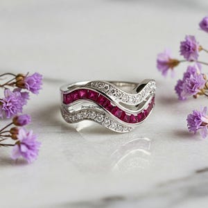 Può includere: Un anello d'argento con un design a onda che presenta due file di pietre di rubino rosso e una fila di pietre di zircone cubico trasparenti. L'anello è su una superficie di marmo bianco.