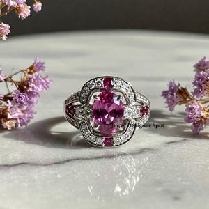 Pode incluir: Um anel de prata com uma grande pedra oval rosa cercada por pedras brancas menores e pedras vermelhas de destaque. O anel é engastado em um design delicado e intrincado.