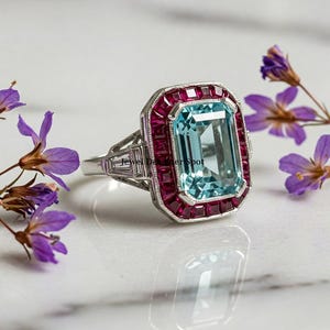 Puede incluir: Un anillo de plata con una gran piedra preciosa de aguamarina de talla esmeralda rodeada por un halo de rubíes rojos. El anillo está sobre un fondo blanco.