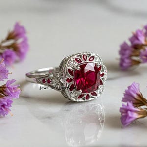 Puede incluir: Un anillo de plata con una gran piedra de rubí de forma cuadrada rodeada de diamantes y rubíes más pequeños. El anillo tiene un diseño delicado de inspiración vintage.