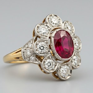 May include: A red ruby and diamond ring set in white and yellow gold. The ruby is oval shaped and surrounded by a halo of round diamonds. The ring has a floral design.