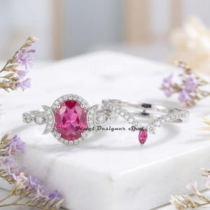 Puede incluir: Juego de anillos de dos piezas con una banda plateada. El anillo principal tiene una piedra rosa de talla ovalada, rodeada de piedras más pequeñas. El conjunto incluye una banda curva con una piedra rosa en forma de marquesa.