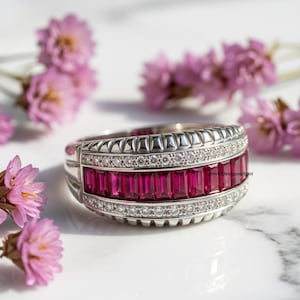 Pode incluir: Um anel de prata com uma faixa de pedras preciosas retangulares vermelho-escuras. O anel é adornado com uma fileira de pequenas pedras preciosas transparentes e uma borda texturizada. Flores cor-de-rosa ao fundo.