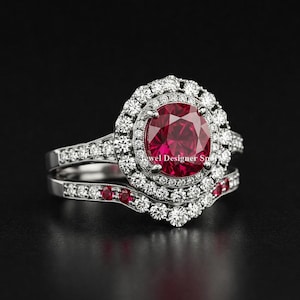 May include: A two-piece engagement ring set with a large, round, ruby-colored center stone surrounded by a halo of smaller diamonds. The band is also adorned with diamonds and smaller ruby-colored stones. The ring is set against a black background.