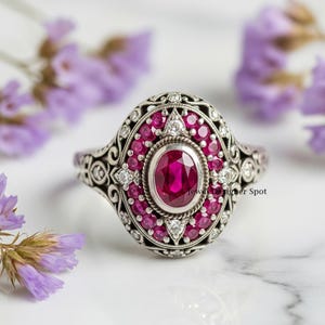 Op de afbeelding: Een sierlijke zilveren ring met een grote ovale robijn in het midden, omringd door kleinere robijnen en accentdiamanten. De ring heeft een ingewikkeld filigraanontwerp.
