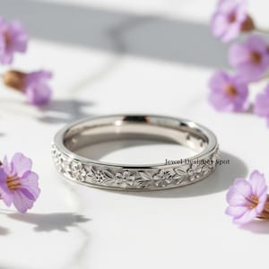 Puede incluir: Un anillo plateado con un diseño floral. El anillo presenta una banda de flores y hojas detalladas. El anillo está sobre un fondo blanco con flores moradas en primer plano. El texto "Jewel Designer Spot" es visible en el anillo.