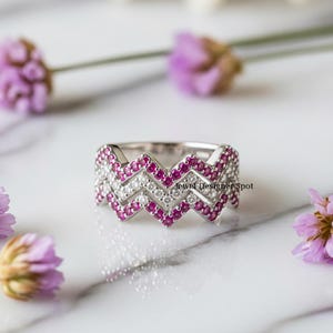 Può includere: Un anello d'argento con un design a zig-zag, incastonato con piccole pietre preziose rotonde rosa e bianche. L'anello è su una superficie di marmo bianco, con fiori viola sfocati sullo sfondo. Il testo "Jewel Designer Spot" è visibile.