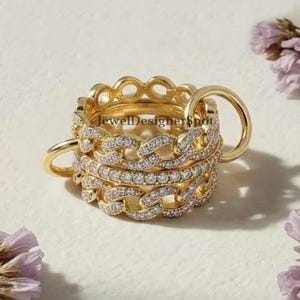 May include: Gold-toned rings with a chain link design, some encrusted with small, clear gemstones. The rings are stacked together, with a simple band ring on the side. The background is a soft, neutral color with a few purple flowers.