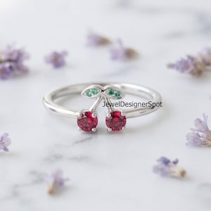 May include: A silver ring featuring a cherry motif with two round red stones as the fruit and two small green stones set into the leaves. The text JewelDesignerSpot is visible on the band. The ring has a polished finish and a thin band.