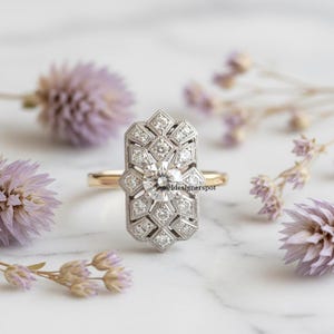 May include: An Art Deco style ring featuring a central round diamond and smaller diamonds in a geometric design. The ring has a gold band and a silver-coloured setting. The ring is displayed on a white surface with purple flowers.