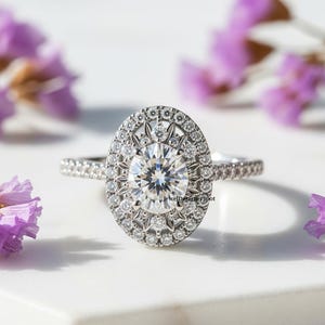 Puede incluir: Anillo de compromiso de diamantes con una gran piedra central de forma ovalada rodeada por un halo de diamantes más pequeños. La banda también está incrustada con pequeños diamantes. Sobre una superficie blanca con flores moradas.