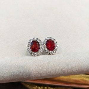 Może przedstawiać: Para owalnych kolczyków rubinowych osadzonych w białym złocie z halo mniejszych diamentów.