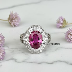 May include: A silver ring with a large, oval-cut, magenta gemstone as the focal point. The gemstone is surrounded by smaller, rectangular, clear stones. The ring has an ornate design with filigree details and is set against a marble background.
