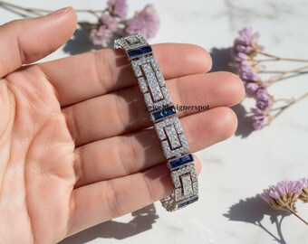 May include: A silver bracelet with a geometric design, featuring rectangular and square sections. The bracelet is adorned with clear and blue gemstones, creating a sparkling effect. The jewelry is held in a hand, with a blurred floral background.