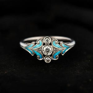 May include: A silver ring with a large, round, clear diamond surrounded by smaller diamonds. The ring is decorated with blue enamel in a swirling, floral pattern.