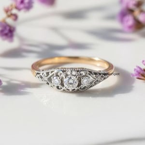 May include: A gold and silver ring featuring three clear gemstones. The ring has an ornate, filigree design around the gemstones. The ring is set against a white background with purple flowers in the background.