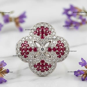 May include: A silver-toned ring with a four-leaf clover design. The ring is adorned with small, round, deep red gemstones and clear, round gemstones. The ring is set against a white background with purple flowers.