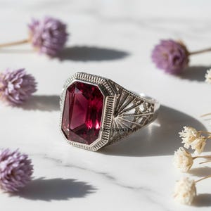 Op de afbeelding: Een zilveren ring met een grote, rechthoekige, dieprode edelsteen. De ring heeft een sierlijk filigraanontwerp rond de edelsteen en op de band. De ring ligt op een wit marmeren oppervlak met gedroogde bloemen.