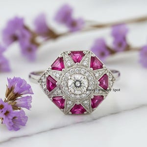 May include: A silver ring with a geometric design. The ring features a central round diamond surrounded by smaller diamonds and triangular pink gemstones. The ring is set against a blurred background of purple flowers.