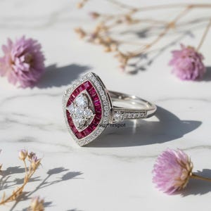 Puede incluir: Un anillo de plata con una cara en forma de marquesa. El anillo presenta un grupo central de pequeños diamantes redondos rodeado por un borde de piedras preciosas rojas y un halo de diamantes más pequeños. La banda está detallada con pequeños diamantes redondos. El texto "jewel designerspot" es visible.