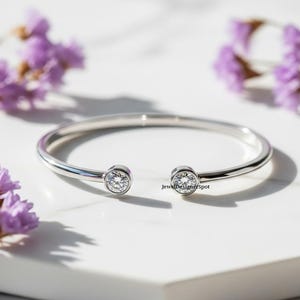 Könnte beinhalten: Ein silbernes, offenes Armband mit zwei runden, klaren Edelsteinen. Das Armband liegt auf einer weißen Oberfläche, mit lila Blumen im Hintergrund. Der Text "JewelDesignerSpot" ist sichtbar.