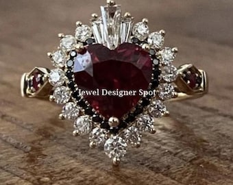 Könnte beinhalten: Ein goldener Ring mit einem großen roten herzförmigen Edelstein, umgeben von kleineren weißen und schwarzen Edelsteinen. Der Ring ist mit einem Heiligenschein aus kleineren weißen Edelsteinen und einer Band aus schwarzen Edelsteinen besetzt. Der Text "Jewel Designer Spot" ist auf dem Ring sichtbar.