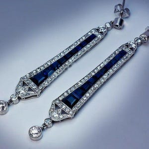 Op de afbeelding: Een paar oorbellen in Art Deco-stijl. Elke oorbel heeft een lang, rechthoekig ontwerp met een rij vierkante, diepblauwe stenen in het midden, omlijst door kleinere, heldere stenen. Onderaan bungelt een ronde, heldere steen.