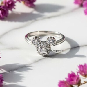 May include: A silver ring featuring a Mickey Mouse design. The ring has a round cluster of clear stones for the face and two smaller stones for the ears. The ring is set against a white marble surface with purple flowers.