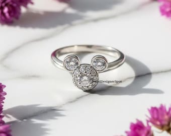 May include: A silver ring featuring a Mickey Mouse design. The ring has a round cluster of clear stones for the face and two smaller stones for the ears. The ring is set against a white marble surface with purple flowers.