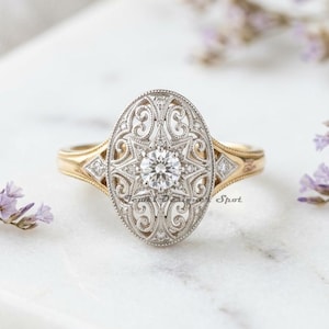 May include: An antique-style ring with a central diamond and an oval, filigree design. The ring features a gold band and a silver-coloured filigree setting, with small accent diamonds. The ring is set against a white background with purple flowers.