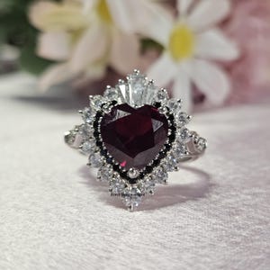 May include: A silver ring with a large, heart-shaped ruby surrounded by smaller white and black diamonds. The ring is set in a delicate, floral design.