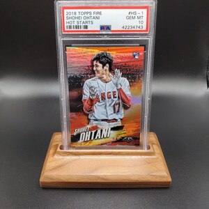May include: A graded baseball card featuring Shohei Ohtani in a red and white baseball uniform with the text "Shohei Ohtani" and "Angels" on the card. The card is graded GEM MT 10 and has a #HS-1 designation. The card is in a clear plastic case and is displayed on a wooden stand.