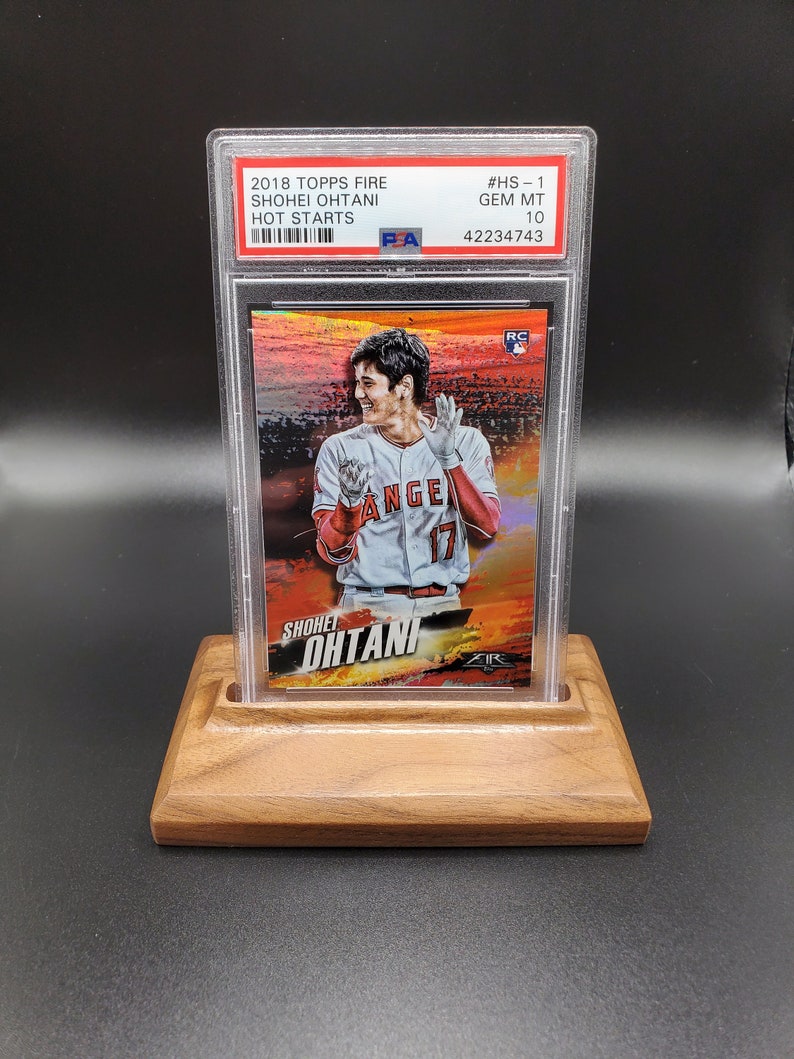 May include: A graded baseball card featuring Shohei Ohtani in a red and white baseball uniform with the text "Shohei Ohtani" and "Angels" on the card. The card is graded GEM MT 10 and has a #HS-1 designation. The card is in a clear plastic case and is displayed on a wooden stand.