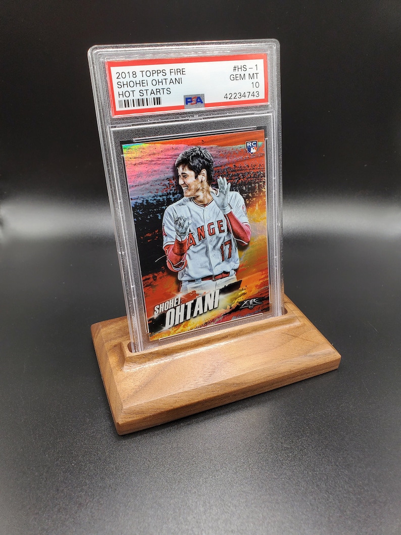 May include: A baseball card featuring Shohei Ohtani in a red and white baseball uniform. The card is graded GEM MT 10 and is from the 2018 Topps Fire Hot Starts series. The card is in a protective case and is displayed on a wooden stand.