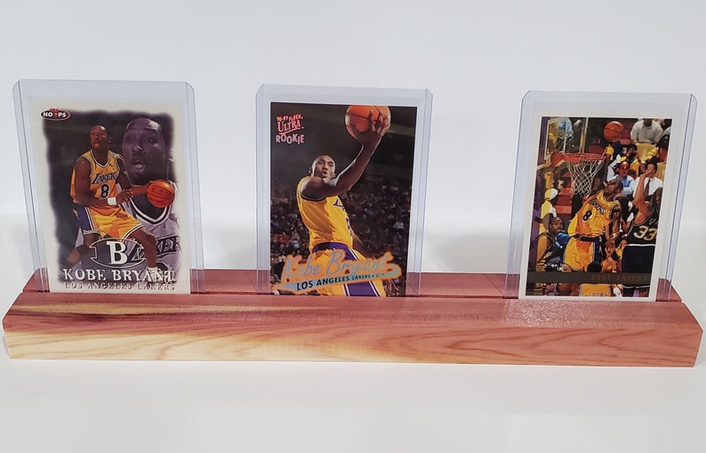 May include: Three Kobe Bryant basketball trading cards in protective plastic cases, displayed on a wooden stand. The cards feature images of Bryant in his Los Angeles Lakers uniform.