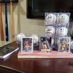May include: Three clear plastic display cases with signed baseballs and three clear plastic display cases with basketball trading cards featuring Kobe Bryant. The display cases are on a wooden stand.